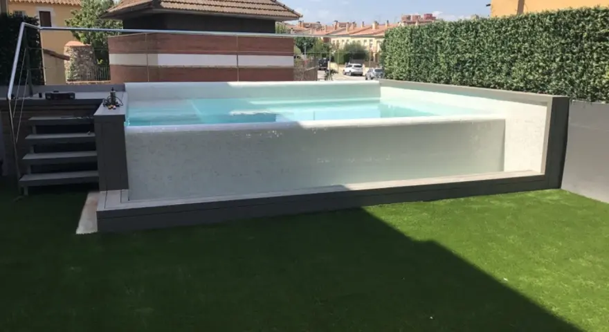 Moderna piscina con mosaico blanco, rodeada de césped artificial y setos. Ideal para decoración de terrazas con piscina.