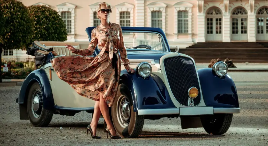 Elegancka kobieta w kwiecistej sukience i okularach przeciwsłonecznych opiera się o zabytkowy samochód. Stylizacja nawiązuje do mody lat 20.