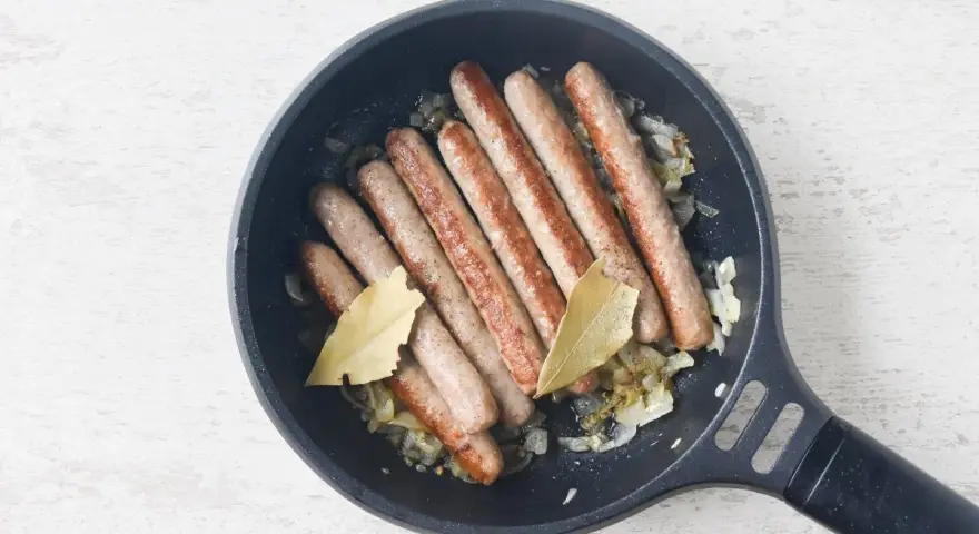 Salchichas doradas cocinándose en sartén con cebolla y hojas de laurel.