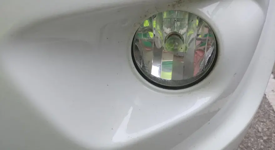 Zbliżenie na biały zderzak samochodu z widocznym okrągłym reflektorem. To światła przeciwmgielne przednie, lekko zakurzone, gotowe do jazdy w trudnych warunkach.