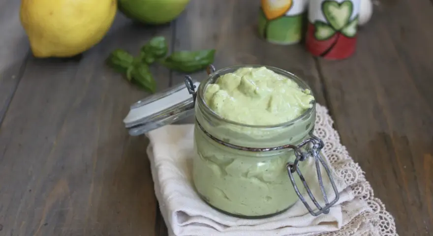 Un vasetto di cremoso pesto al limone, con limoni e basilico sullo sfondo.