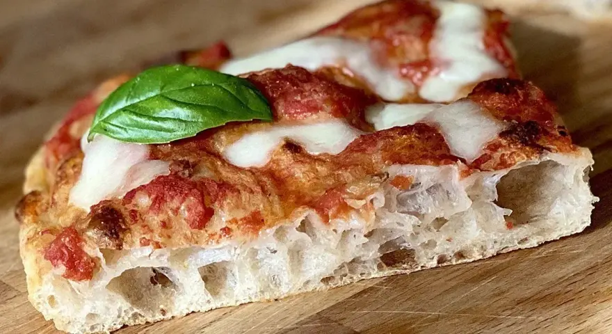 Fetta di pizza fatta in casa teglia, con crosta alveolata, pomodoro, mozzarella filante e una foglia di basilico fresco.