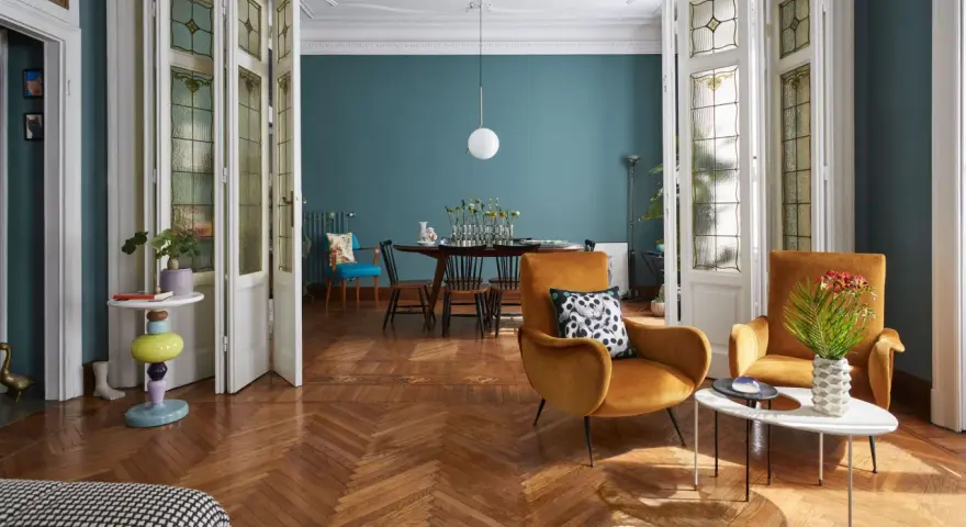 Salotto con un mix di arredamento moderno e antico: poltrone ocra, tavolino colorato, porte a vetri d'epoca e un tavolo da pranzo in stile classico.
