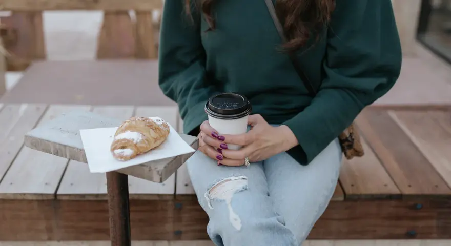 Une femme en pull vert, jean déchiré, savoure un café et un croissant. Parfait pour un look décontracté.