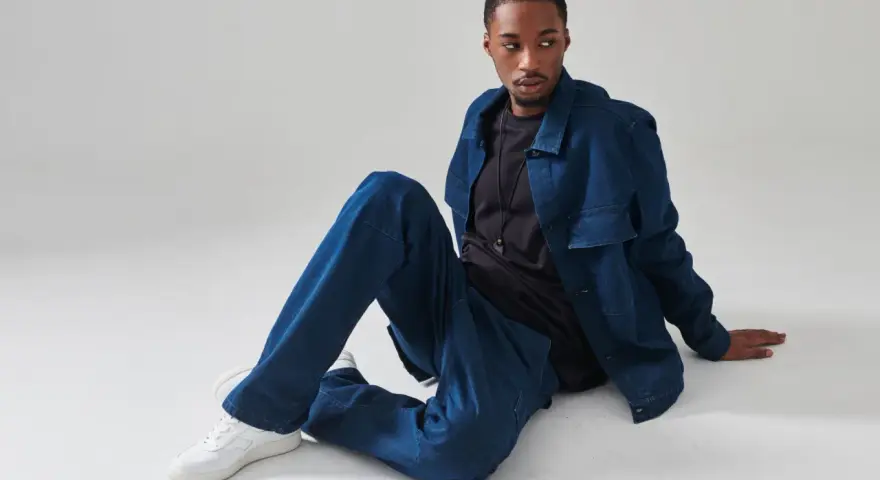 Giovane uomo con completo in materiale jeans, seduto a terra, con giacca e pantaloni oversize.