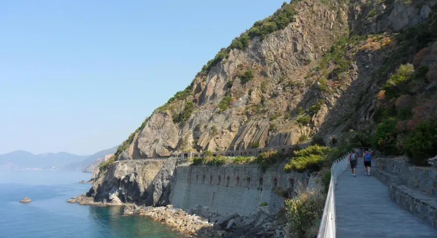 Wanderer auf dem Sentiero Azzurro entlang der felsigen Küste Liguriens. Das Meer glitzert türkisblau neben dem steilen Hang.
