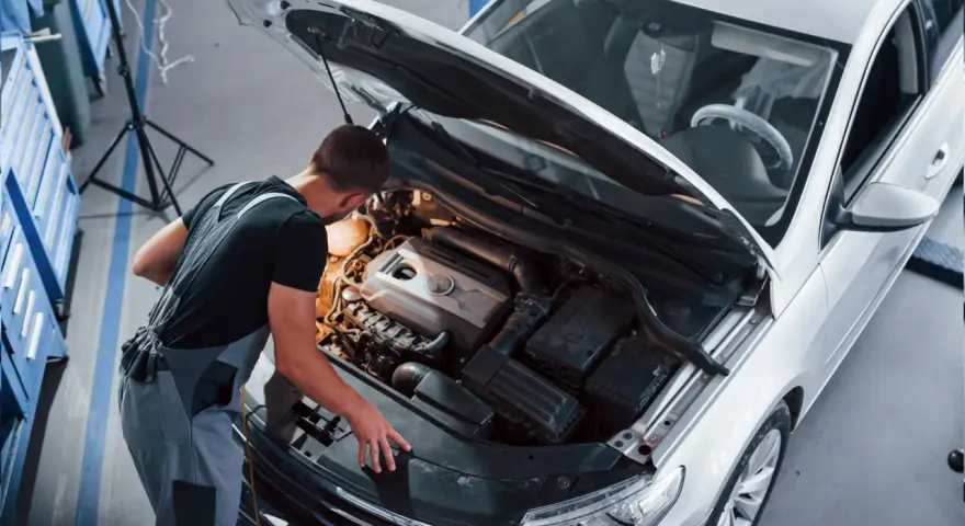 Młody mechanik w kombinezonie pracuje przy silniku białego samochodu. To jego pasja, każdy konstruktor marzy o takiej precyzji.