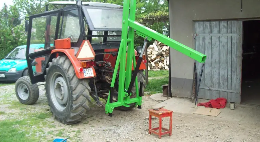 Czerwony traktor z zamontowanym zielonym ładowaczem TUZ przed budynkiem gospodarczym.