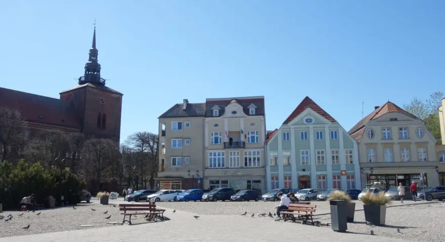 Słupsk, stary rynek: kościół z wieżą, kolorowe kamienice, ławki, ludzie i gołębie na brukowanym placu.