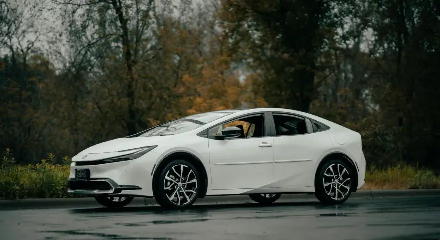 Biała Toyota Prius zaparkowana na mokrej drodze, z drzewami w tle.