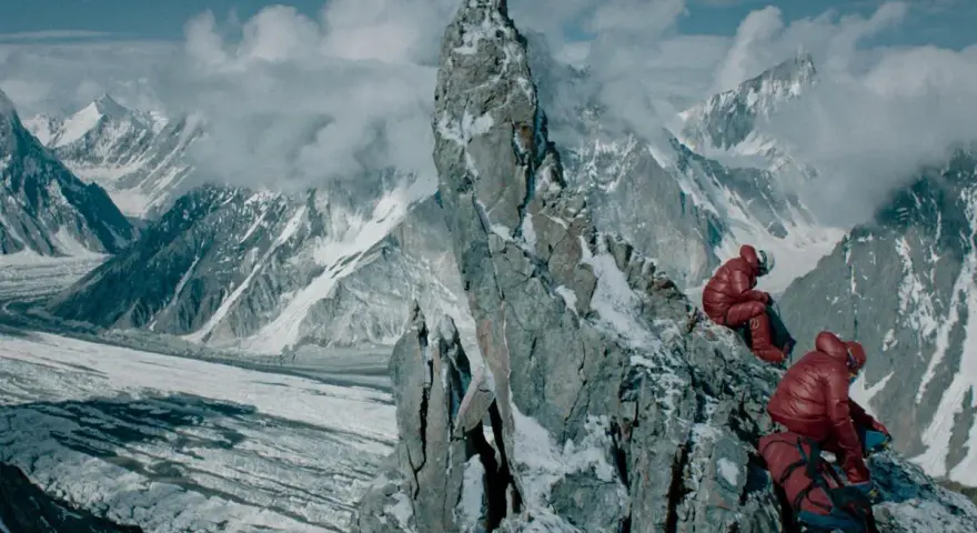 Dwóch wspinaczy w czerwonych kurtkach odpoczywa na skalistej grani. W tle rozciąga się lodowiec i ośnieżone szczyty. To obraz **broad peak tragedia**.