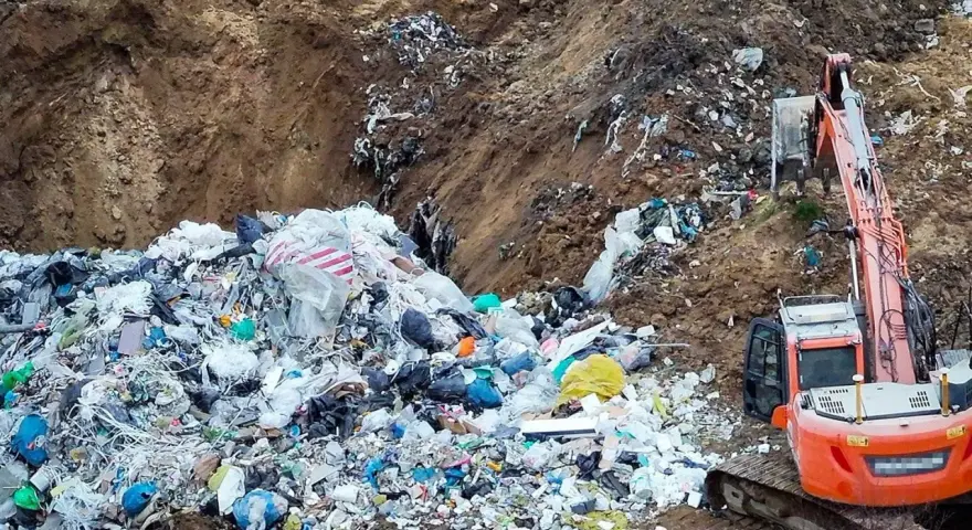 Ogromna góra śmieci, tworząca nielegalne wysypisko, z koparką w tle. Widać worki, folie i różne odpady.