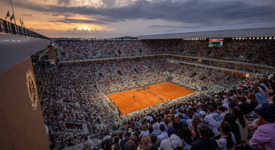 Widok na pełny stadion podczas meczu na kortach ziemnych. Zachodzące słońce oświetla trybuny pełne widzów, tworząc atmosferę emocjonującego **Roland Garros live**.