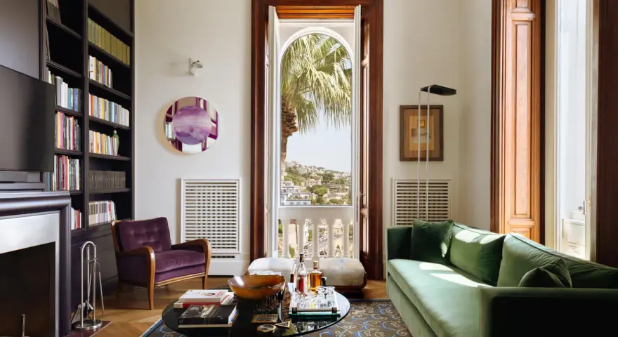 Soggiorno elegante in una casa stile Liberty, con divano verde, poltrona viola, libreria e vista panoramica.