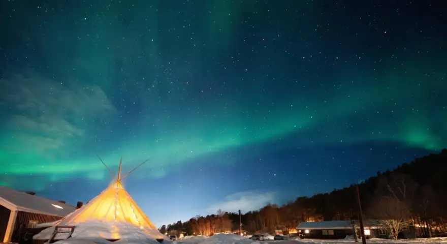 Nocne niebo nad ośnieżoną wioską, gdzie nie zachodzi słońce. Tańcząca zorza polarna nad oświetlonym tipi i domkami.