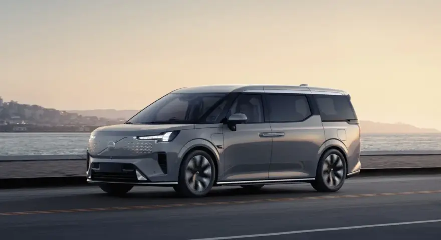 Szary Volvo EM90 jedzie wzdłuż wybrzeża o zachodzie słońca.