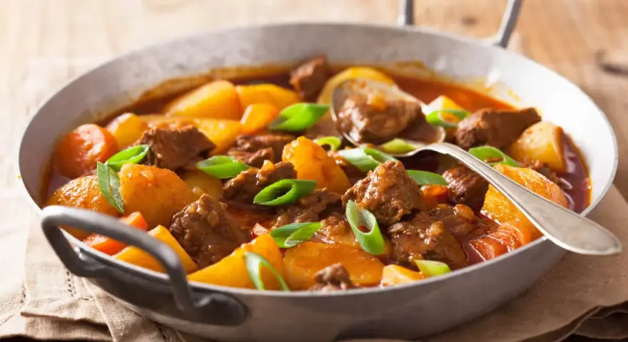 Estofado de ternera tierno con patatas y zanahorias en una sartén, adornado con cebolleta.