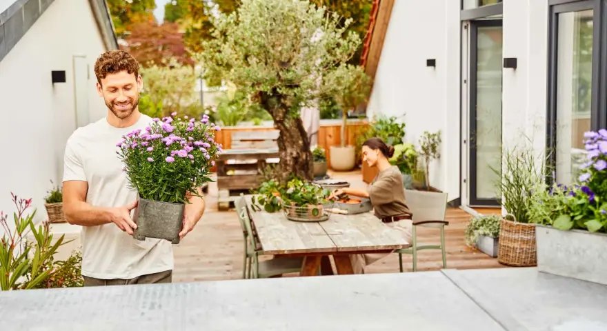Mann hält Topf mit lila Blumen, bereit, die Dachterrasse zu bepflanzen. Eine Frau sitzt am Tisch und arbeitet im Garten.