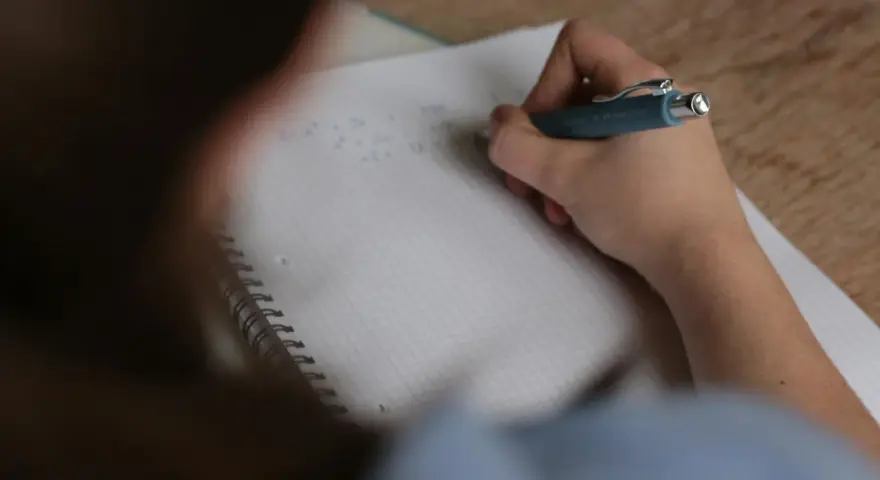 Uczeń pisze w zeszycie, zastanawiając się, ile trwa egzamin ósmoklasisty.
