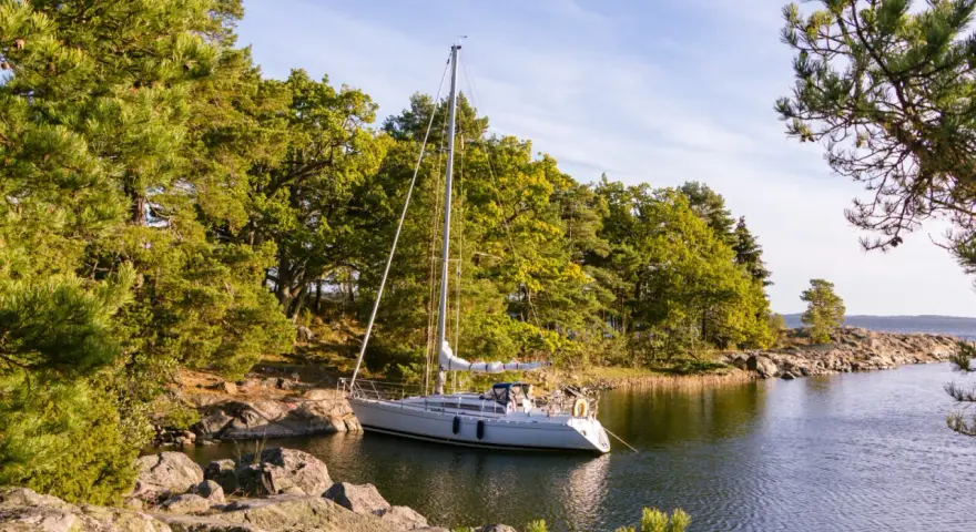 Segelboot liegt ruhig im Wasser, bereit, den Anker zu legen oder zu setzen. Grüne Bäume und Felsen säumen die Küste.