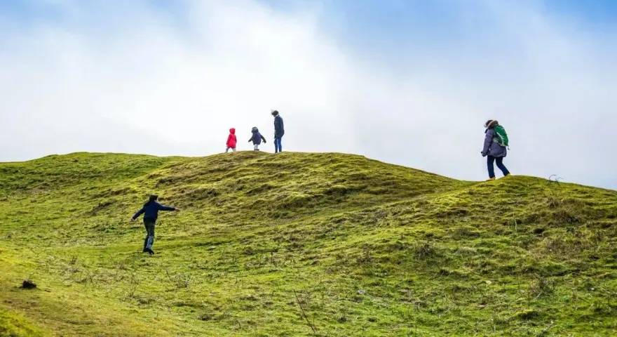Famiglia esplora sentieri per bambini su una collina verdeggiante sotto un cielo azzurro e nuvoloso.