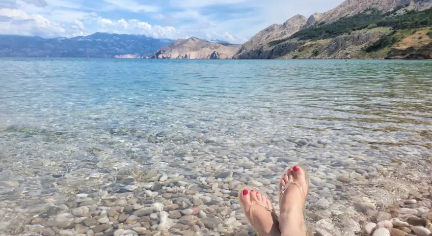 Stopy w klapkach na kamienistej plaży w **Krk National Park**. Woda krystalicznie czysta, w tle góry.