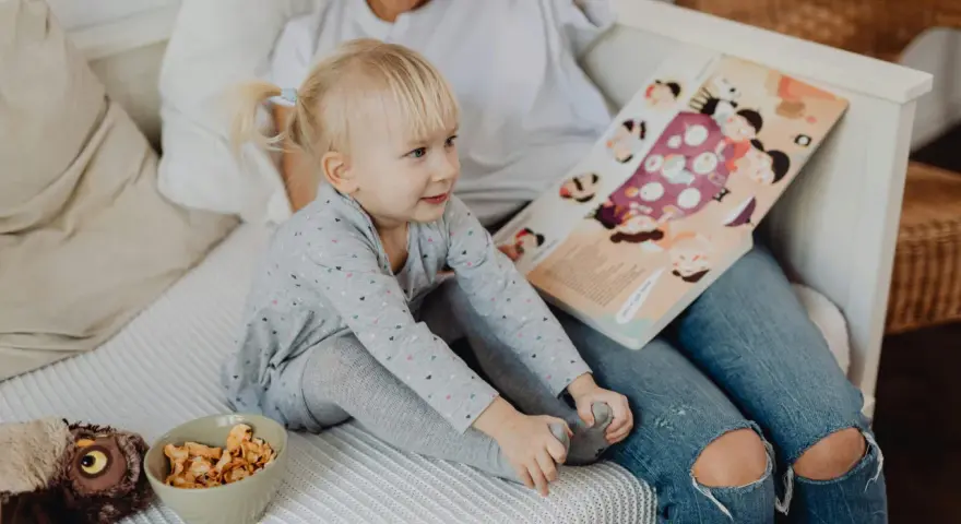 Mała dziewczynka z blond kucykami słucha bajki czytanej przez opiekunkę. Zastanawiasz się, ile zarabia opiekunka dla dziecka?