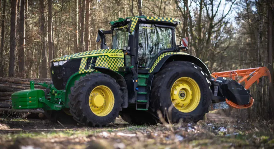 Zielony traktor John Deere z pomarańczowym mulczerem, gotowy do pracy w lesie.