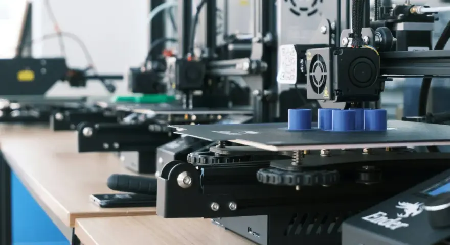 Drukarki 3D pracują, tworząc niebieskie cylindry. W tle widać kolejne maszyny, a na biurku leży smartfon, gotowy do zarządzania drukiem przez Raspberry Pi z OctoPrint.