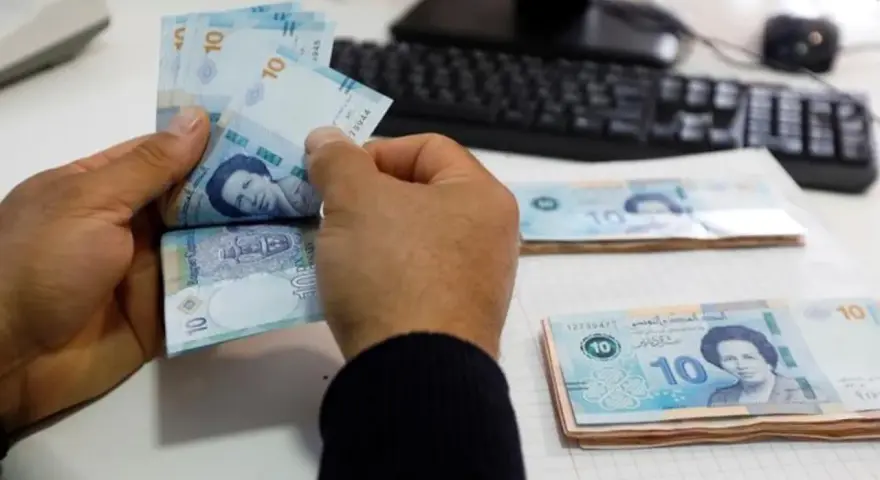Ręce liczą banknoty 10 dinarów, tunezyjska waluta. Na stole leżą stosy pieniędzy, a w tle widać klawiaturę komputera.