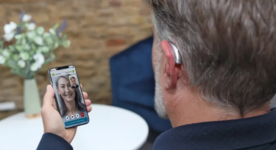 Mężczyzna z aparatem słuchowym rozmawia przez wideorozmowę na telefonie. Aplikacja na telefon pozwala na zdalne dopasowanie aparatów.
