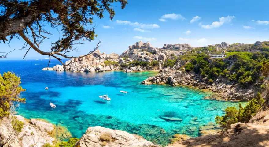 Turkusowa woda i skaliste wybrzeże Costa Smeralda na Sardynii. Kilka łodzi kołysze się na falach.