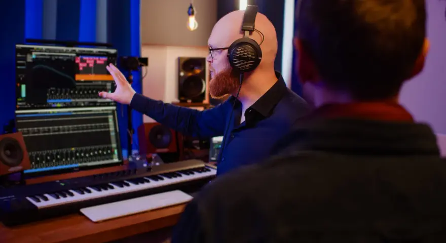Tontechniker mit Kopfhörern erklärt seinem Kollegen am Mischpult die Einstellungen. Er bittet um Feedback zur Abmischung.