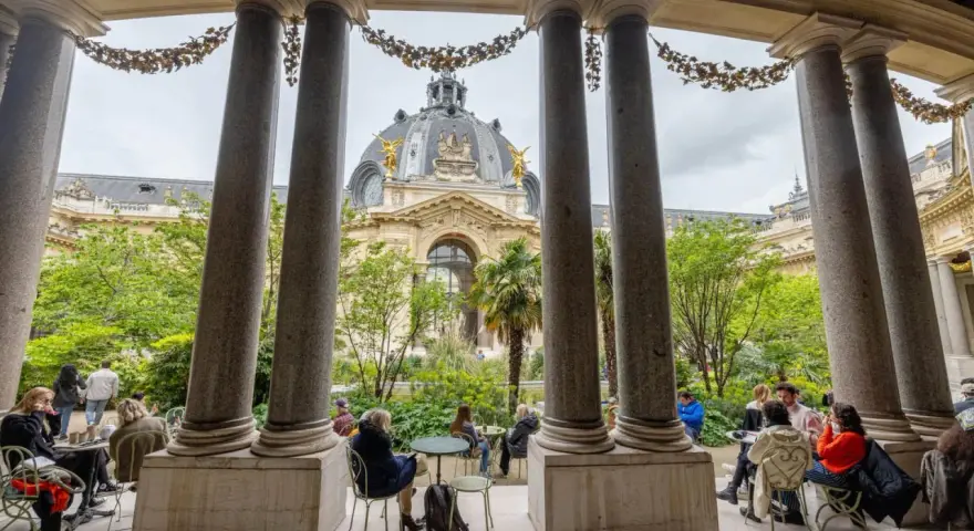 Vue sur le Grand Palais depuis un portique, avec des visiteurs profitant d'un café dans le jardin. Un petit palais, un grand palais.