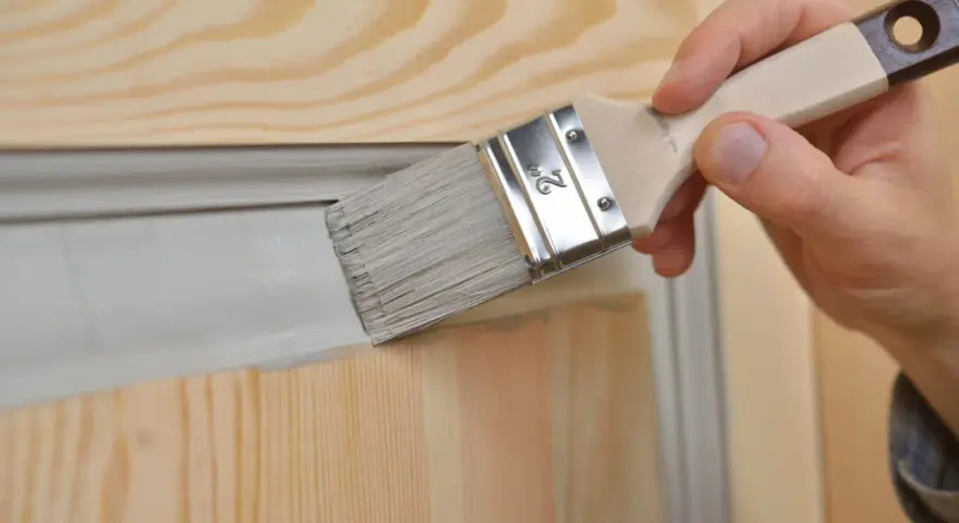 Une main utilise un pinceau pour relooker une porte avec des moulures, appliquant une peinture grise sur le bois clair.
