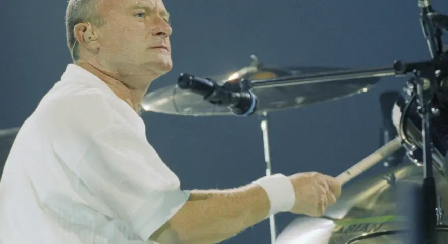 Phil Collins, w białej koszuli, gra na perkusji. Legendarny muzyk, znany z zespołu Genesis.