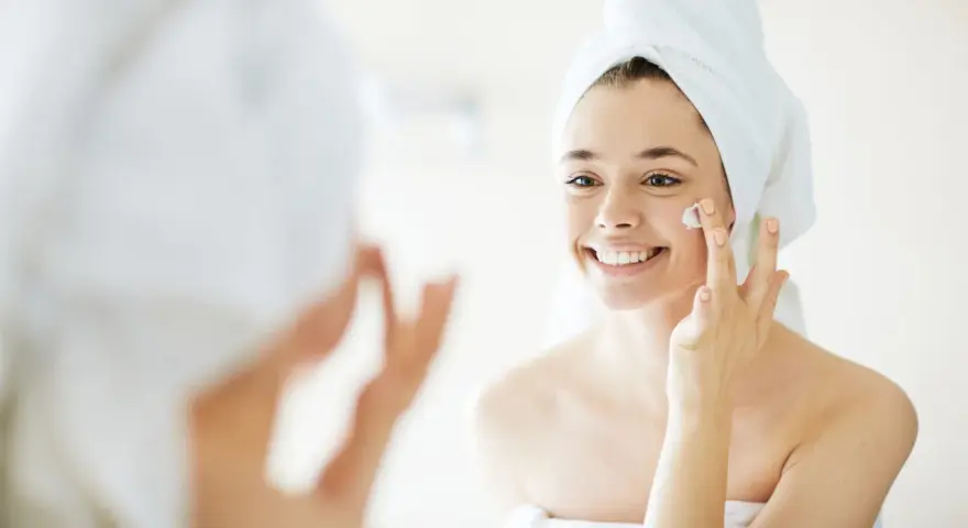 Una mujer sonriente con toalla en la cabeza se aplica crema en la cara, un paso esencial para como cuidar la piel de la cara.