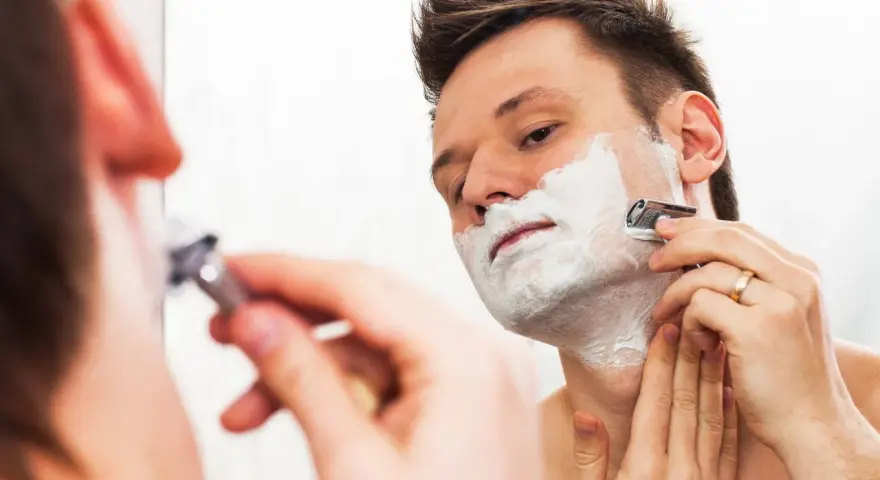 Hombre con espuma de afeitar en la cara, afeitándose con una maquinilla. ¿Es mejor afeitarse antes o después de la ducha?