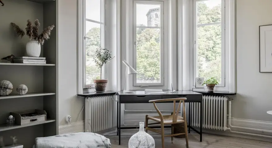 Una **scrivania sotto finestra** con vista sul verde. Un angolo studio accogliente con piante, lampada e una sedia di design.