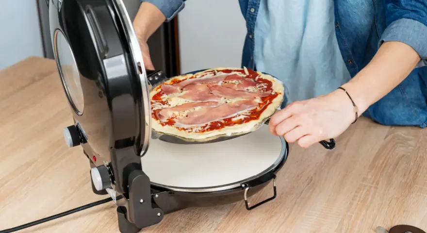 Osoba wkłada surową pizzę z szynką do okrągłego pieca do pizzy.