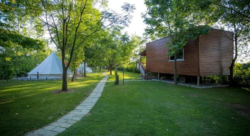 Malowniczy camping w Albanii z białymi namiotami typu bell i drewnianym domkiem na palach, otoczony zielenią.