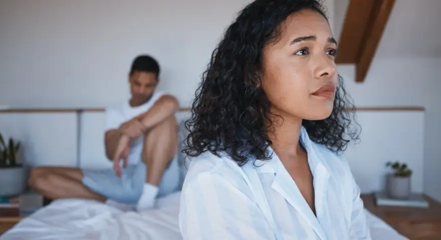 Une femme pensif sur le lit, son partenaire en arrière-plan. Elle pense : il est gentil mais je m'ennuie.