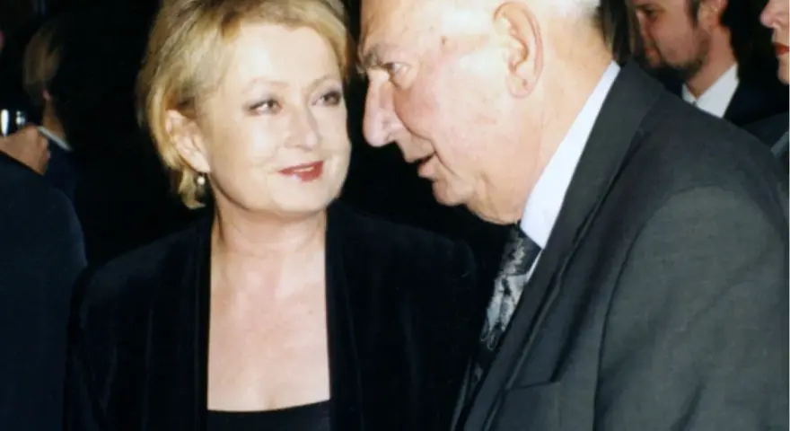 Ewa Holoubek w czarnej sukni i marynarce rozmawia z mężczyzną w garniturze.