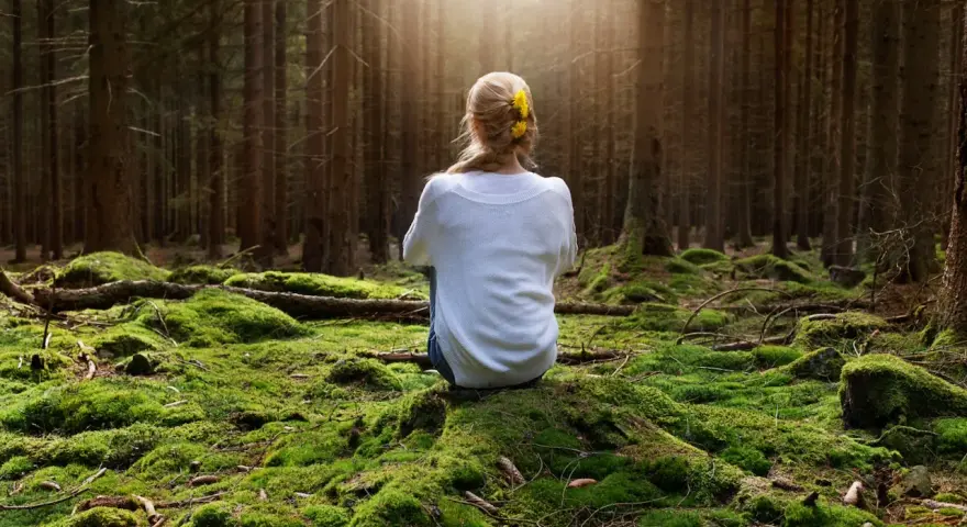 Frau sitzt im Wald auf moosbewachsenem Boden, umgeben von Bäumen. Sonnenstrahlen fallen durch das Blätterdach und erzeugen eine Atmosphäre der Katharsis.