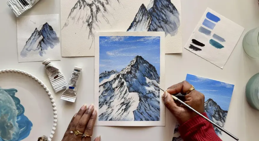 Une main peint une peinture montagne facile, avec des tubes de peinture et des nuanciers bleus à proximité.