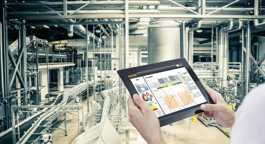 Operator kontroluje linię produkcyjną za pomocą tabletu, na którym widnieje wykres zużycia energii. W tle widać skomplikowany system rur i taśmociągów. Czujnik podczerwieni może być kluczowy dla monitorowania procesów.