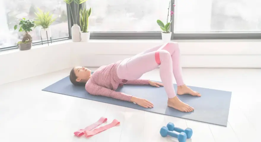 Femme faisant un pont avec une bande de résistance pour comment muscler les fessiers. Haltères et bandes roses à proximité.
