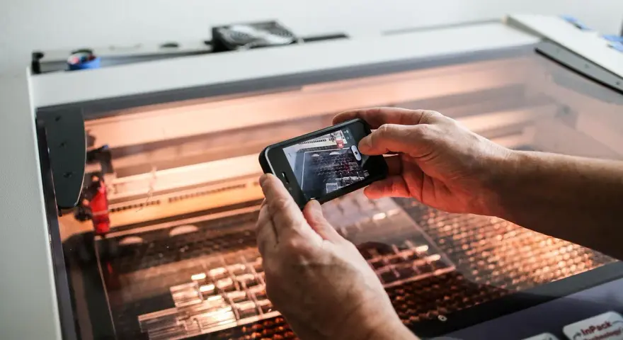 Hands hold a phone, capturing a laser cutter working on intricate acrylic projects.