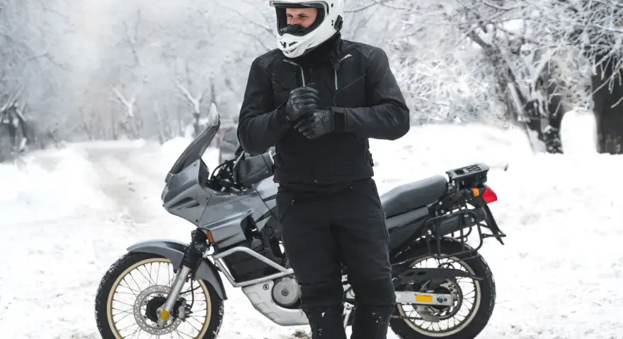 Motociclista con casco bianco e abbigliamento invernale nero, pronto per un'avventura in moto inverno su una strada innevata.