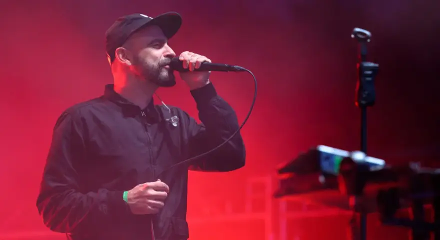 Wokalista z brodą i czapką na głowie śpiewa do mikrofonu na scenie. Czerwone światła tworzą atmosferę koncertu. Tauron Nowa Muzyka line-up.